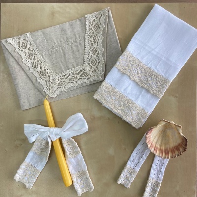 Envelope de tecido bege com renda, toalha branca com rendas, vela amarela com laço rendado e concha do mar com fita rendada