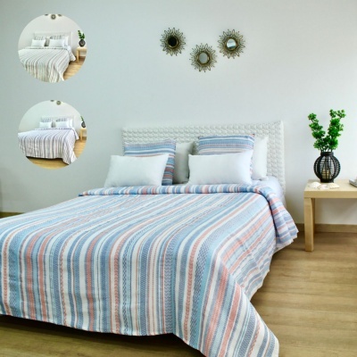 Cama com colcha listrada azul, branco e vermelho claro com almofadas brancas, cabeceira trançada branca e mesa de cabeceira com vaso e flores