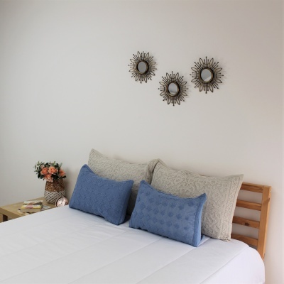 Cama com almofadas azuis e bege, mesa com jarra de flores, três espelhos decorativos na parede branca.