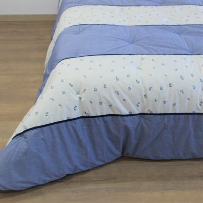 Edredão azul e branco com padrão floral e riscas sobre chão de madeira