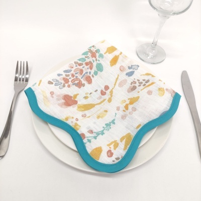 Guardanapo de tecido branco com estampas coloridas e borda azul turquesa numa mesa de refeição