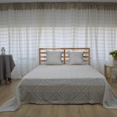 Cama com colcha bege e almofadas, cortinas brancas, duas mesas com objetos decorativos