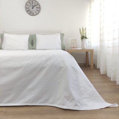 Cama branca com colcha e almofadas, mesa de cabeceira com flores e relógio na parede.