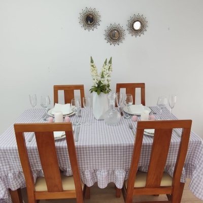 Mesa de jantar com toalha xadrez lilás e branco, cadeiras de madeira, copos, pratos brancos, vaso com flores e espelhos na parede branca