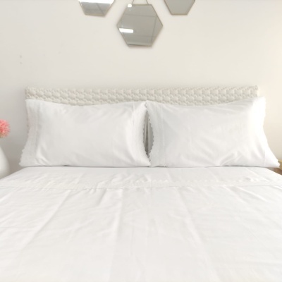 Conjunto de cama branco com fronhas e lençol numa cama com cabeceira trançada e parede branca com espelhos hexagonais