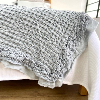 Manta cinzenta texturizada com borda rendada em sofá branco em interior com chão de madeira.