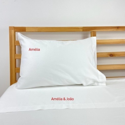 Travesseiro e lençol bordados em branco com nomes vermelhos numa cama com cabeceira de madeira