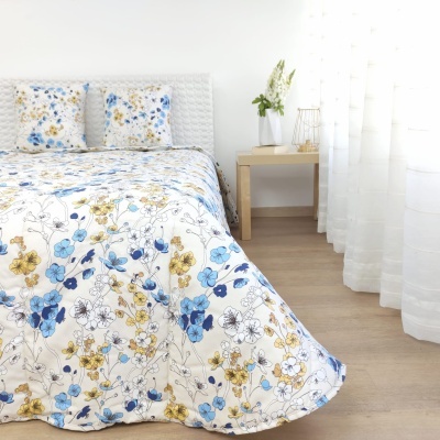 Cobertor de cama floral azul e amarelo com fronhas combinando em quarto com cabeceira branca e mesa de madeira clara