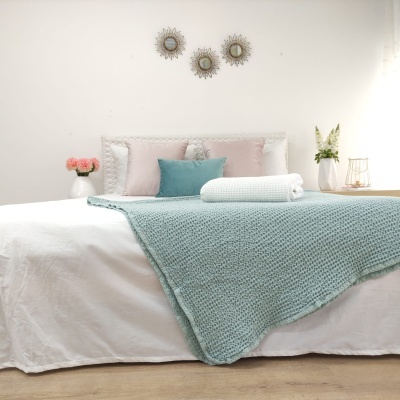 Cama com lençol branco e manta azul claro texturizada com almofadas cor de rosa e azul num quarto decorado
