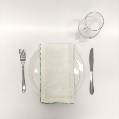 Talheres, prato branco, guardanapo branco com borda verde e copo de vidro sobre superfície branca.