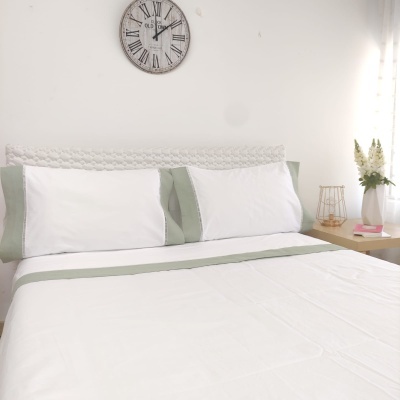 Cama com roupa de cama branca e verde, relógio na parede e mesa com flores