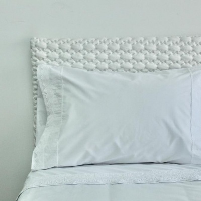Almofada branca com fronha bordada sobre cama branca e cabeceira texturizada
