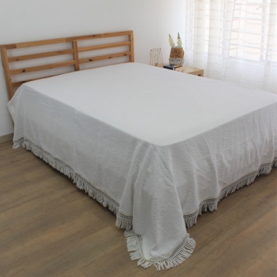 Cama de madeira clara com colcha branca e franjas, junto a mesa de cabeceira e janela com cortina branca