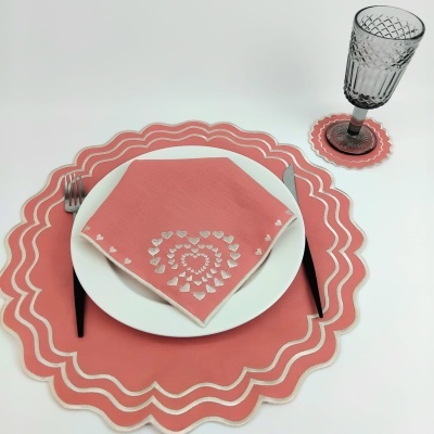 Conjunto de jantar com base de prato, porta-copo e guardanapo rosa com padrões brancos, prato branco, talheres pretos e copo cinzento.