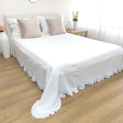 Cama com colcha branca com babados e almofadas, vaso com flores e mesa em ambiente claro.