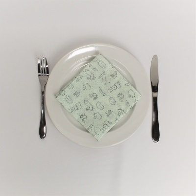Conjunto de mesa com prato branco, talheres de aço inoxidável e guardanapo de tecido verde claro com desenhos pretos.