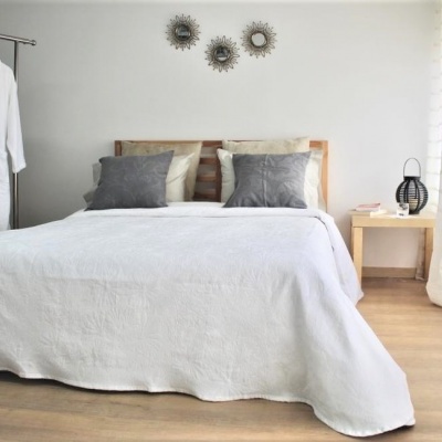 Quarto com cama de madeira, colcha branca, almofadas bege e cinza, mesa de cabeceira com abajur e roupa pendurada