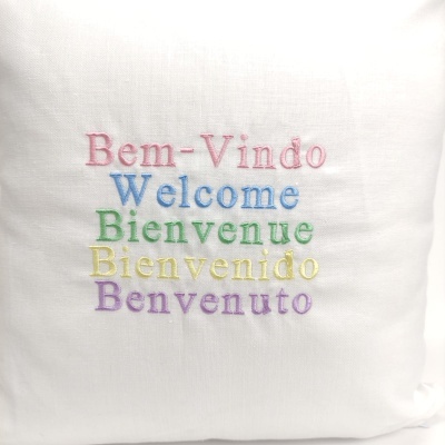 Capa de almofada branca com bordados coloridos das palavras bem-vindo em várias línguas.