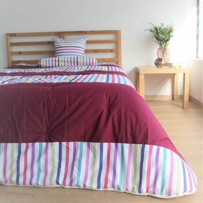 Colcha de cama com riscas coloridas, almofada e rolo, mesa de madeira com vaso de flores