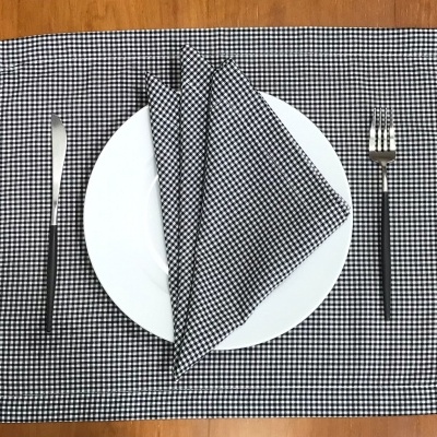 Toalha e guardanapo preto e branco com padrão quadriculado, prato branco, faca e garfo com cabos pretos sobre mesa de madeira