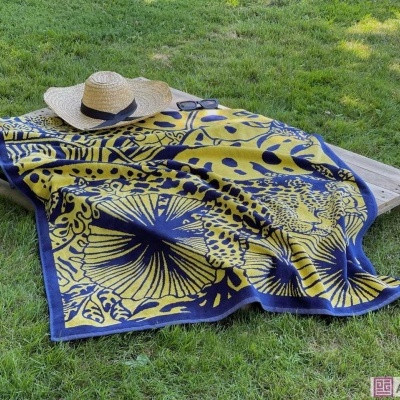 Toalha quadrada azul e amarela com padrão floral, chapéu de palha e óculos sobre estrutura de madeira na relva