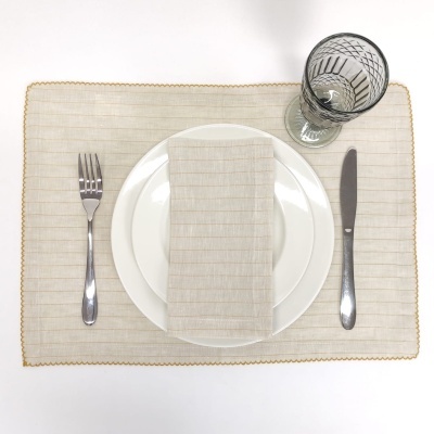 Conjunto de jantar com placemat bege e borda dourada, prato branco, talheres prateados, guardanapo bege riscado e copo de vidro com padrão preto