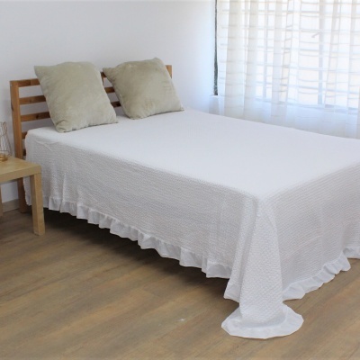 Cama de madeira clara com colcha branca e duas almofadas cinza, perto de janela com cortina branca