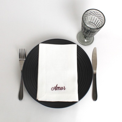 Conjunto de mesa com prato preto, guardanapo branco bordado, talheres e copo de vidro cinzento