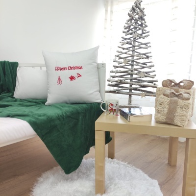 Almofada branca com mensagem de Natal e símbolos em vermelho sobre sofá com manta verde, mesa de madeira clara com caneca, livro e presente decorativo perto de árvore de Natal de paus de madeira