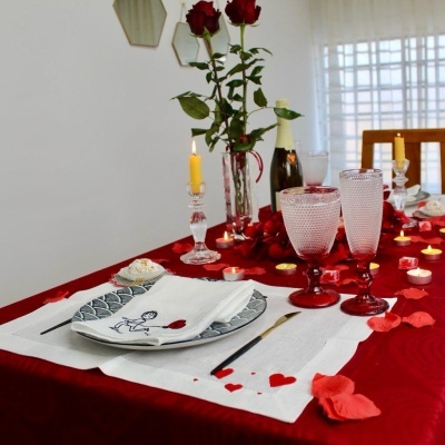 Mesa posta com toalha vermelha, sousplat azul claro, guardanapo branco, castiçais com velas acesas, taças de vidro e pétalas de rosa