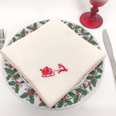 Porta-guardanapos de tecido branco com bordado vermelho sobre prato decorado com folhas de azevinho, garfo, faca e copo vermelho.