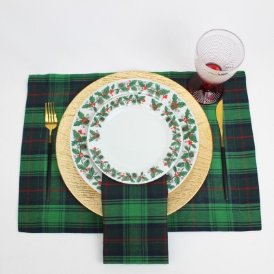 Mesa posta com guardanapo e lugar de mesa tartan verde, pratos decorados com folhas e bagas, copo de vidro com base vermelha e talheres dourados.