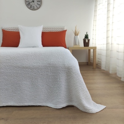 Cama com colcha branca matelassê, almofadas laranja e branca, mesa de cabeceira com vasos, relógio de parede cinzento