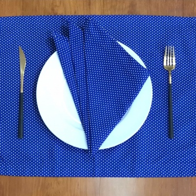Conjunto de mesa com prato branco, individual e guardanapo azuis com bolinhas brancas, talheres dourados com cabo preto e mesa de madeira