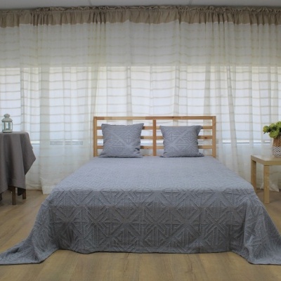 Quarto com cama em madeira clara, colcha e almofadas cinzentas, mesas laterais com decoração, cortinas brancas e chão de madeira