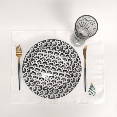 Conjunto de jantar com pratos brancos e pretos, talheres dourados e pretos, copo transparente e toalhete branco com árvore de Natal bordada.