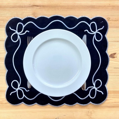 Jogo americano preto com padrão de laços brancos e prato branco numa mesa de madeira