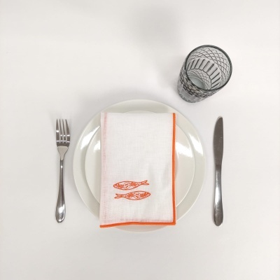 Conjunto de utensílios de mesa com prato branco, guardanapo branco com desenho de peixes laranja, talheres em aço e copo transparente com padrão negro