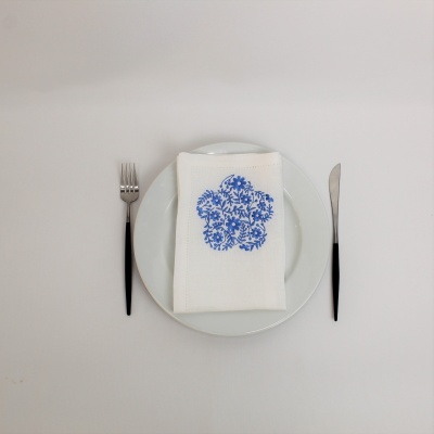 Prato branco com guardanapo branco bordado a azul e talheres com cabo preto num fundo branco