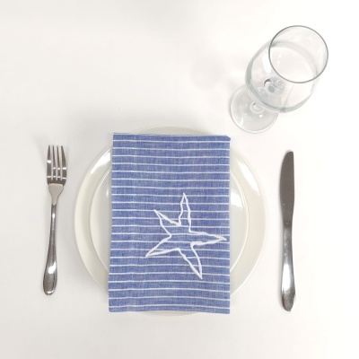Mesa posta com prato branco, guardanapo azul às riscas com estrela, garfo, faca e copo de vidro vazio