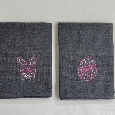 Dois panos de prato cinzentos com bordados rosa de coelho e ovo, em superfície branca.