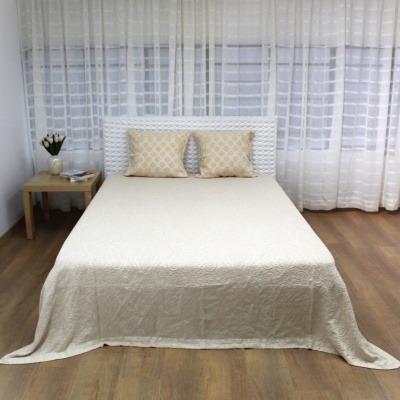 Cama de casal com colcha e almofadas bege, cabeceira branca e mesa de cabeceira em ambiente com cortinas brancas.