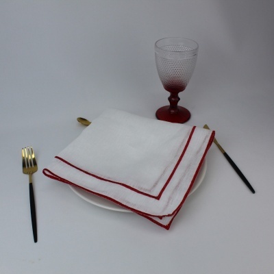 Conjunto de mesa com prato branco, guardanapo branco com borda vermelha, talheres pretos com detalhes dourados e copo transparente com base vermelha.
