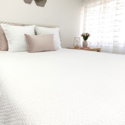 Cama com colcha branca, almofadas brancas e bege claro, mesa de cabeceira com vaso de flores e vela