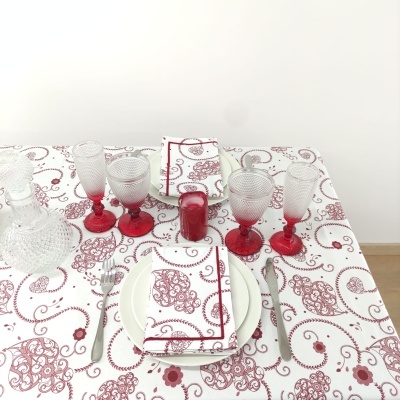 Mesa com toalha branca estampada em vermelho, copos de vidro com haste vermelha, pratos brancos, guardanapos com padrão vermelho, garrafa e vela vermelha.