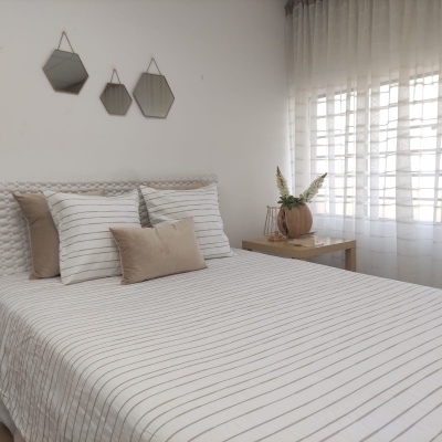 Cama com roupa de cama às riscas e almofadas bege, mesa de madeira com jarro e plantas, cortinas brancas e espelhos hexagonais.