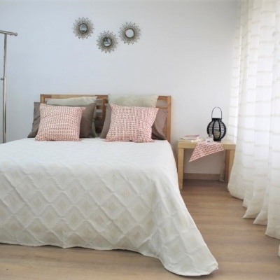 Quarto com cama, colcha branca, almofadas cor de rosa, cortinas brancas e mesa de cabeceira com candeeiro