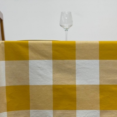 Toalha de mesa com padrão de grandes quadrados em amarelo, bege e branco, com taça de vidro ao fundo