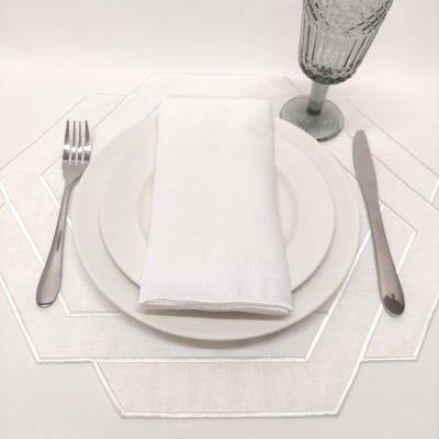 Conjunto de jantar com pratos brancos, talheres metálicos, guardanapo branco, copo cinzento texturizado e jogo americano branco geométrico.