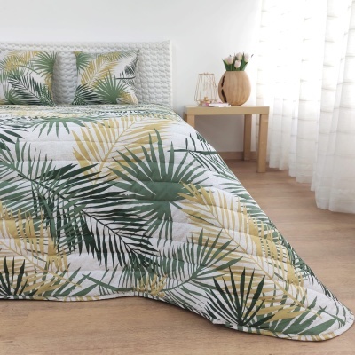 Colcha de cama com padrão de folhas verdes e amarelas, almofadas combinando, mesa de madeira e vaso com flores num quarto iluminado